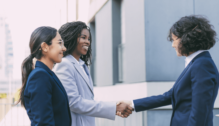 Interracial business partners greeting each other near office building. Business women wearing office suits, shaking hands with each other outside in city. Cooperation concept