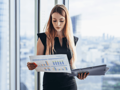 Female financial analyst holding papers studying documents standing against window with city view.