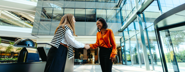 A welcoming scene of two professional women exchanging a friendly handshake in a modern office lobby. The spacious glass surroundings evoke a sense of openness and collaboration.